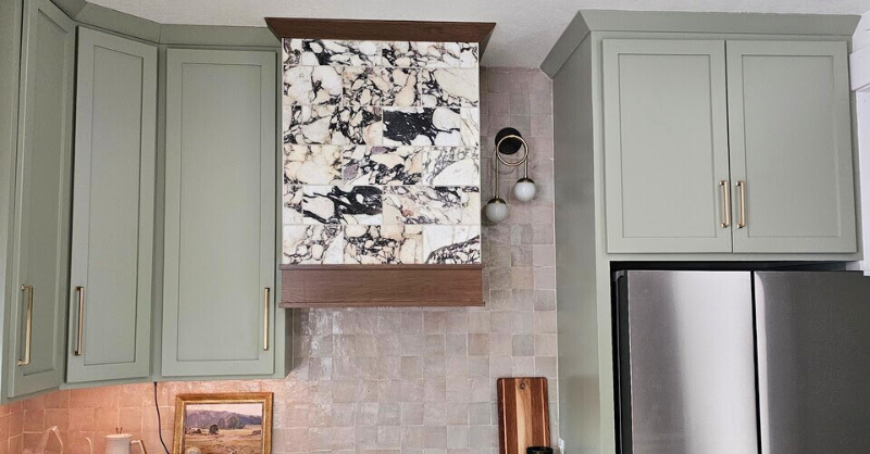 Kitchen with light sage green cabinets with gold handles and a marble oven vent with dark brown trim.