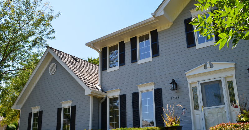 The front of a light blue painted home with dark shutters and bright green plants outside.