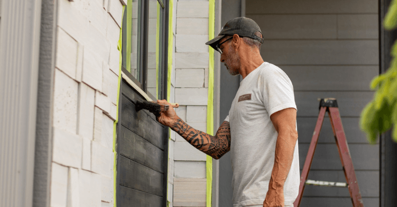 Brush & Roll Painting painter crew lead brushing black paint on exterior side of a home next to window.