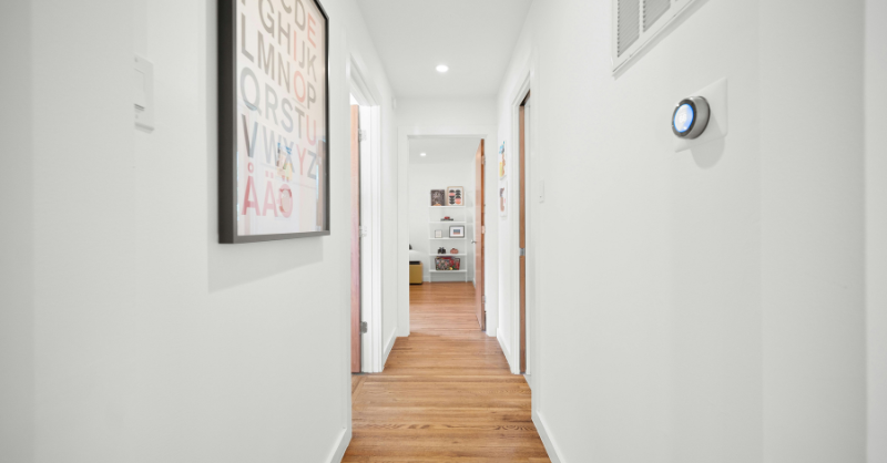 Thermostat hanging on a wall in a hallway with white paint on walls and trim.