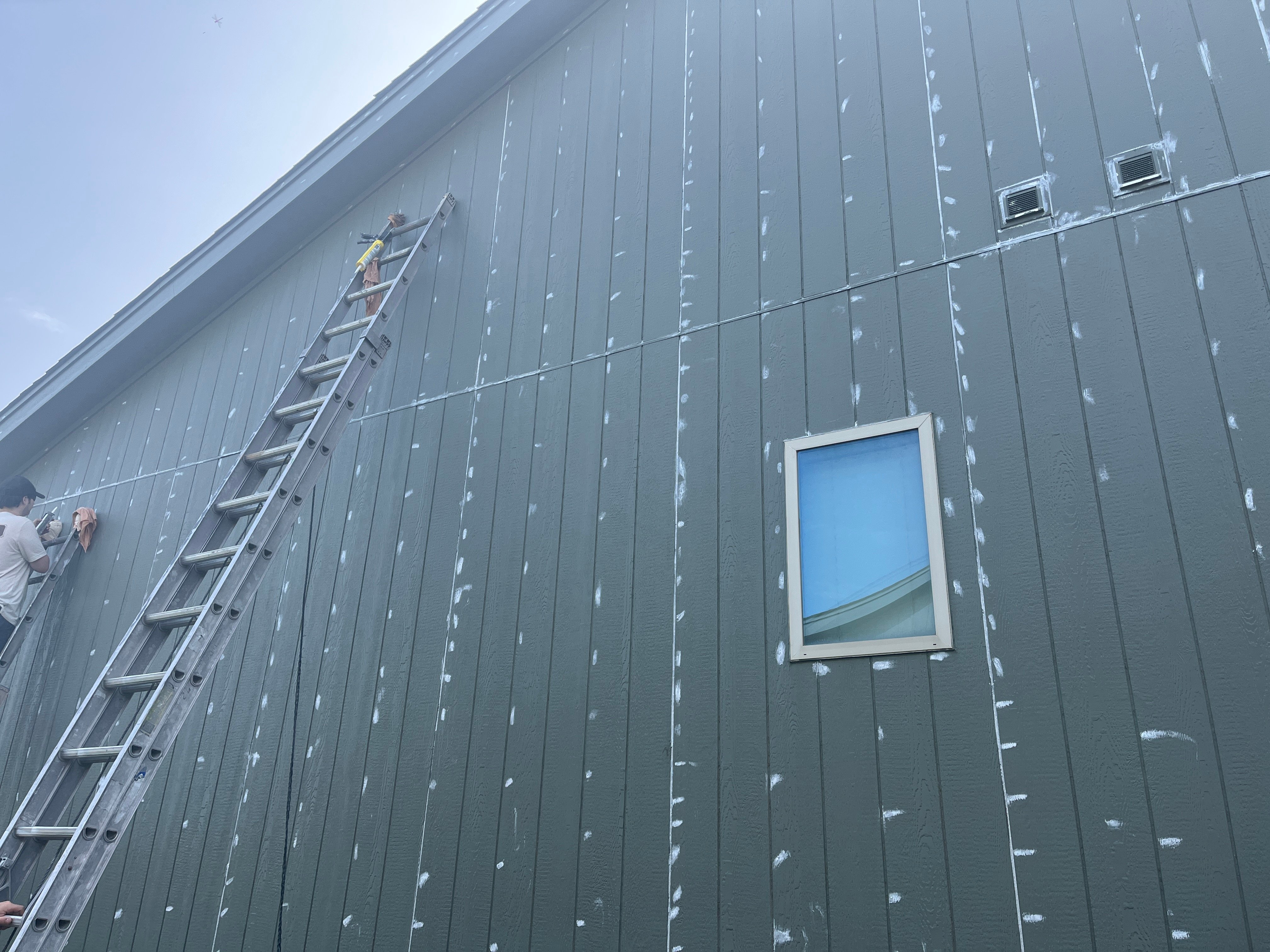 Exterior of home being caulked by a painter. White caulk is along gaps and holes on top of old green paint.